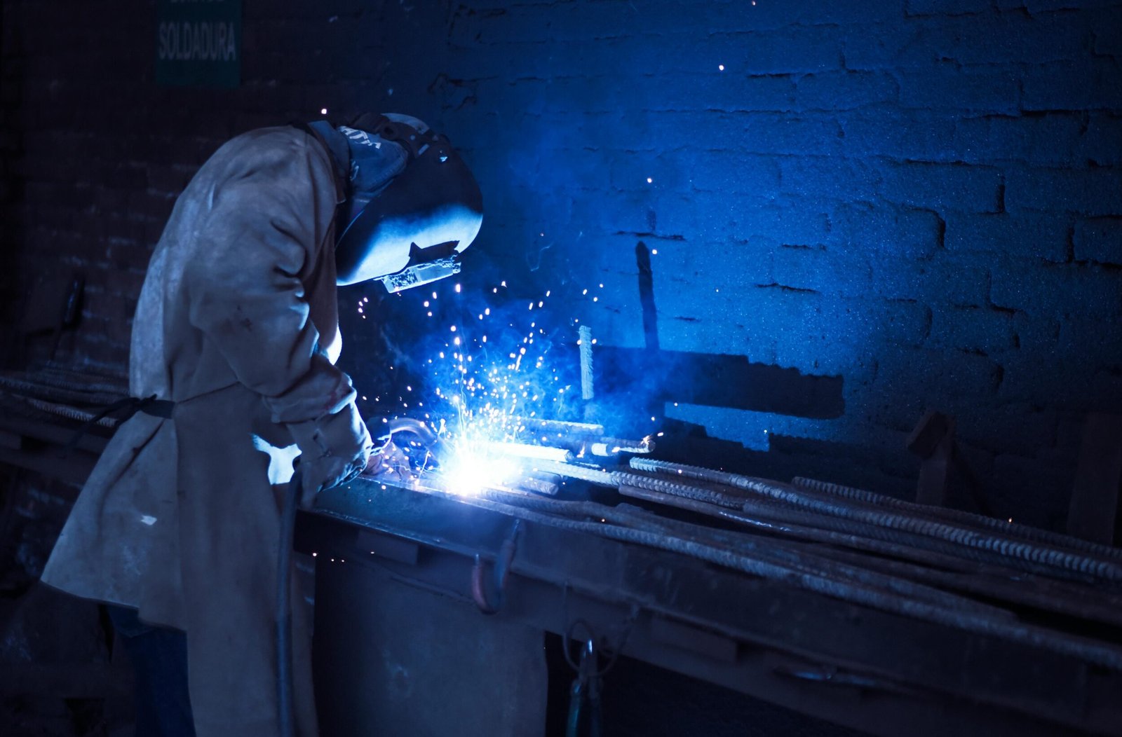 Industrial worker welding metal indoors, emitting bright blue sparks and smoke.