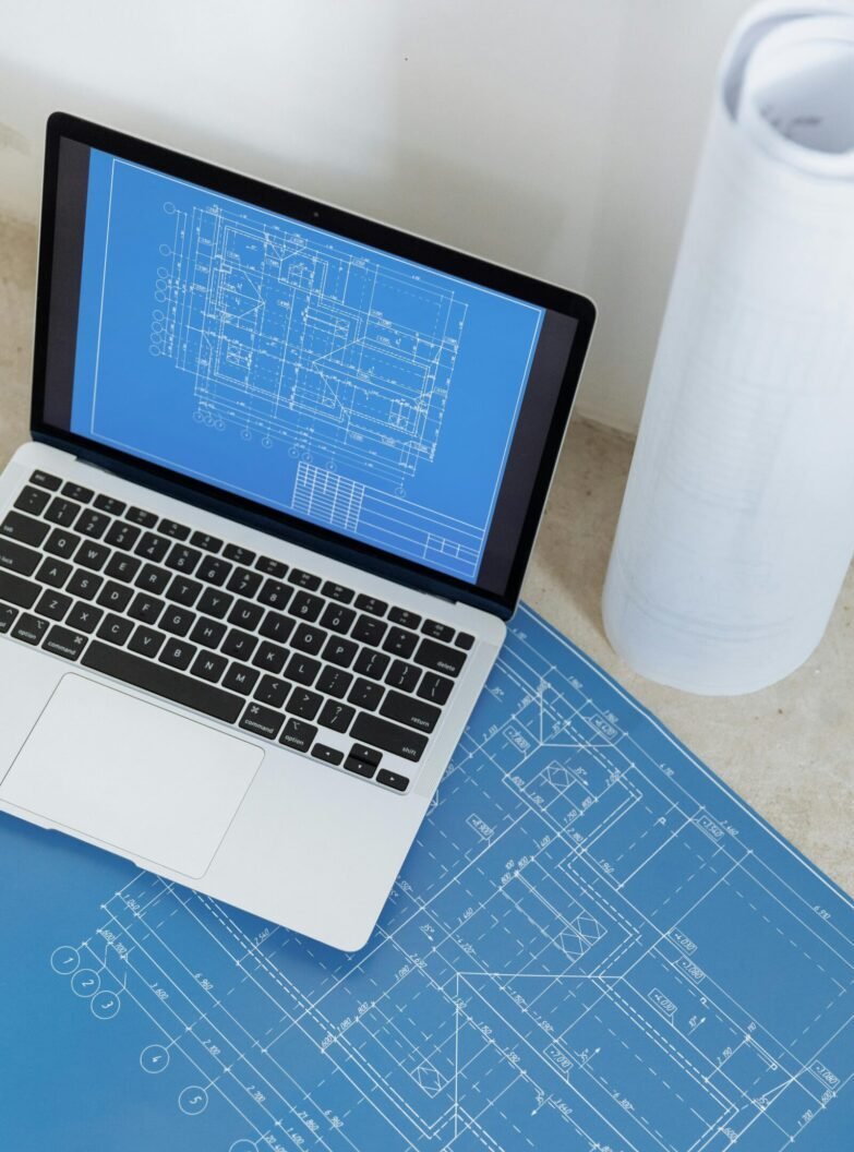 Laptop showing engineering blueprint with rolled paper and drawings on table.